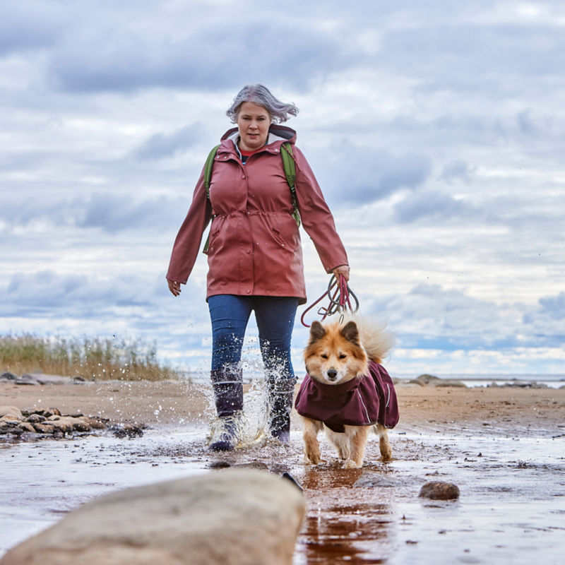 A woman walks her small dog in a winter coat across a wet beach.