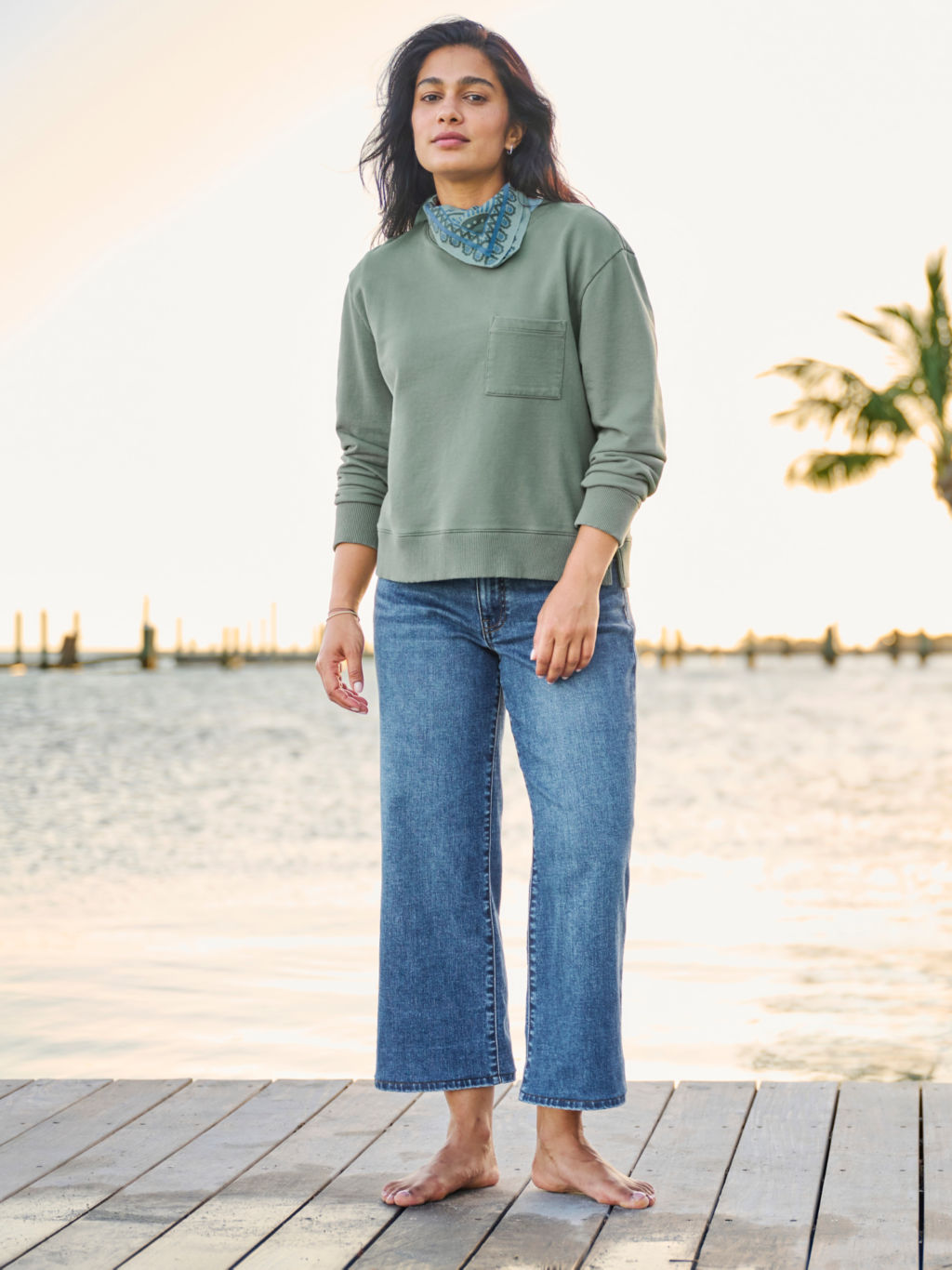 A woman wearing a sage green sweatshirt, wide leg cropped jeans, standing barefoot on a wooden dock.