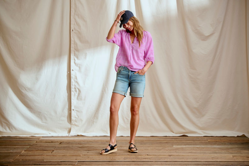 Woman wearing pink shirt and jean shorts in a studio.