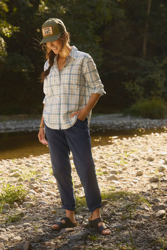 A model wearing an olive trucker hat and a plaid button down shirt with her hand in her pocket.