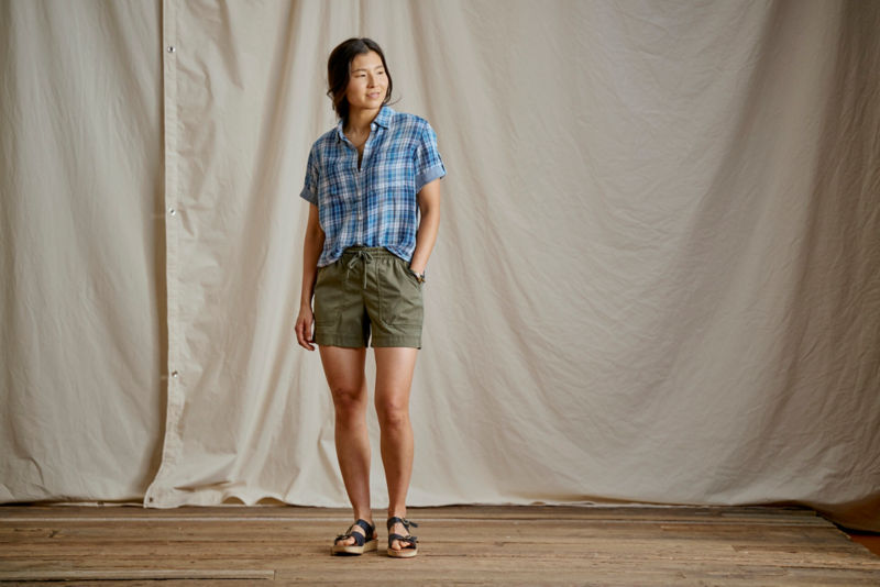 Woman wearing a blue plaid camp shirt and green shorts in a studio.