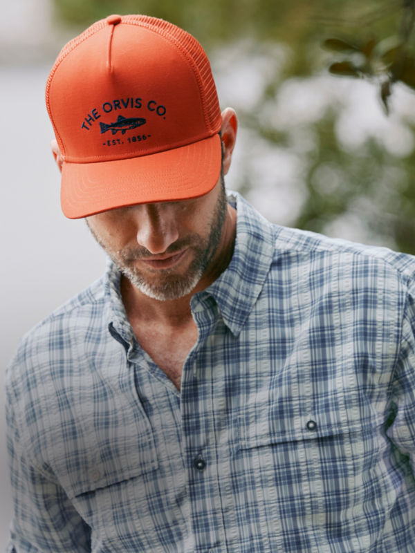 A hiker wearing a blaze orange Orvis ball cap and a blue and white plaid shirt.