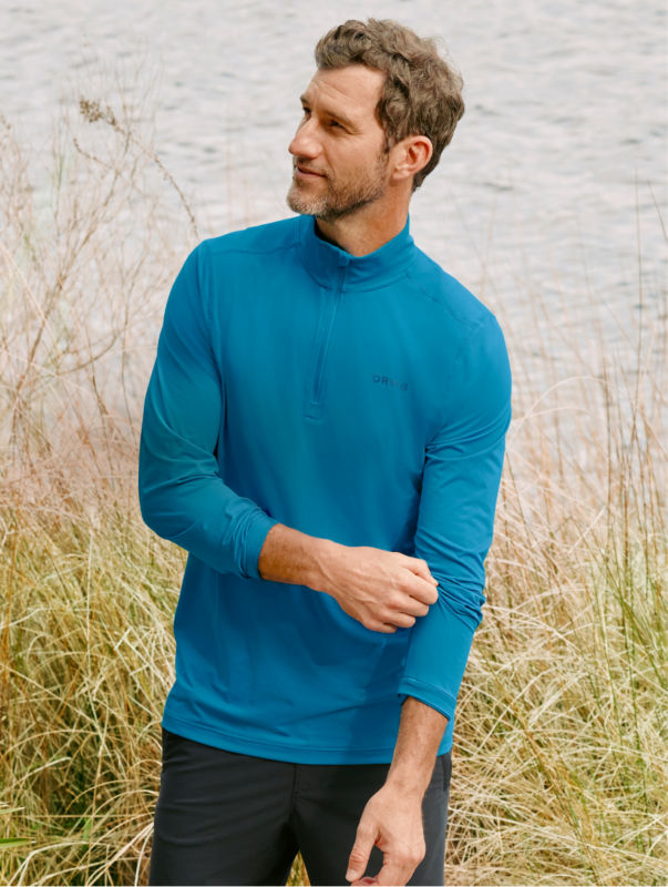 A man in a blue quarter-zip long-sleeved shirt standing in front of water.