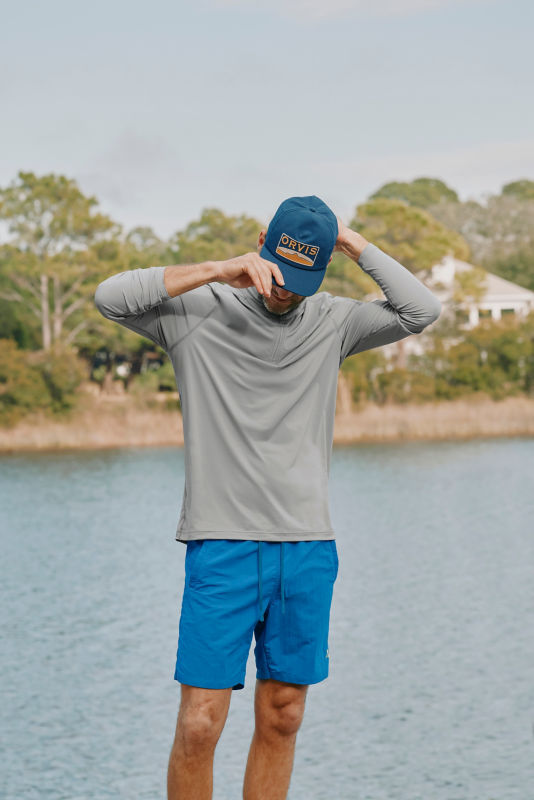 A man putting on an Orvis baseball cap, wearing a long-sleeved grey t-shirt and bright blue shorts.