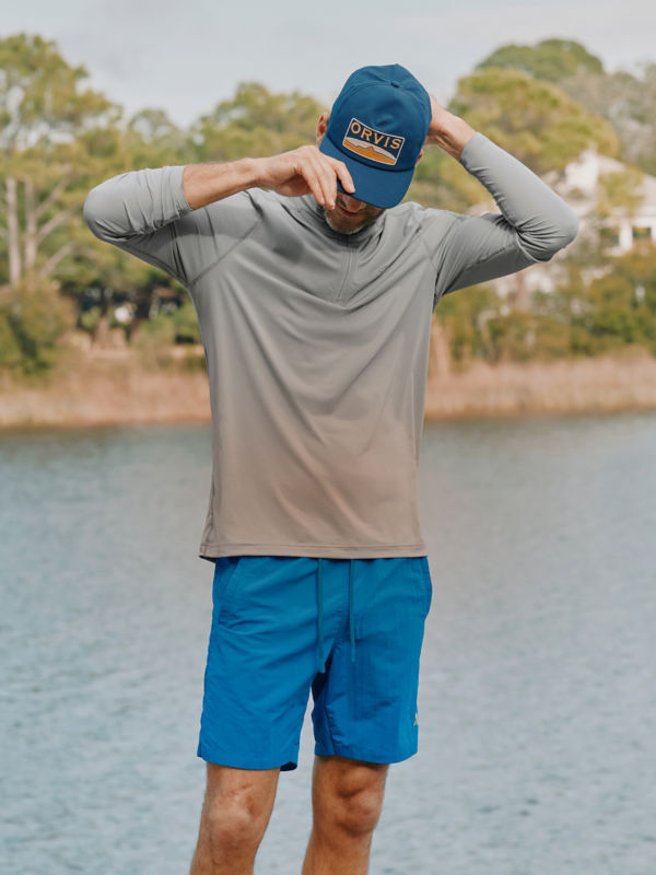 A man wearing a gray sun protective shirt and bright blue pants pauses by the water.