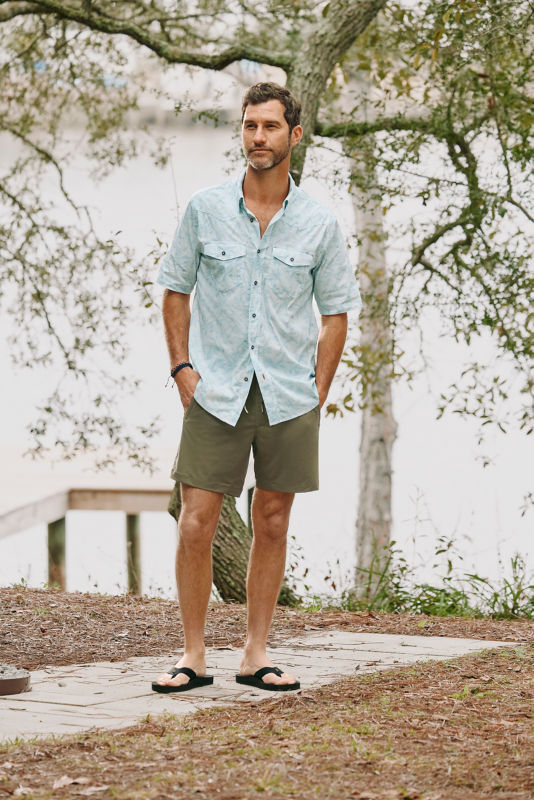Man wearing printed blue shirt and green shorts standing in front of the water.