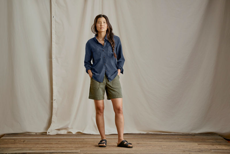 Woman wearing a navy long sleeved camp shirt and green shorts in a studio.