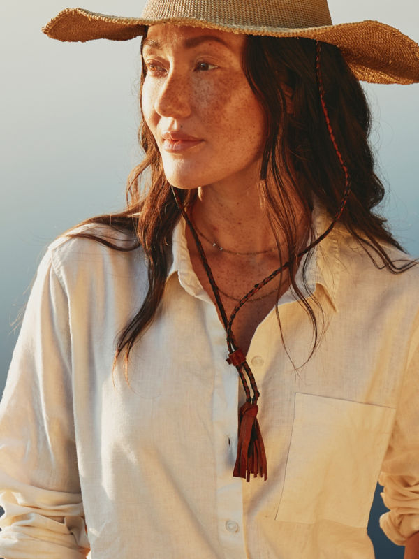 A model stands in front of still water wearing a white linen shirt and straw hat.