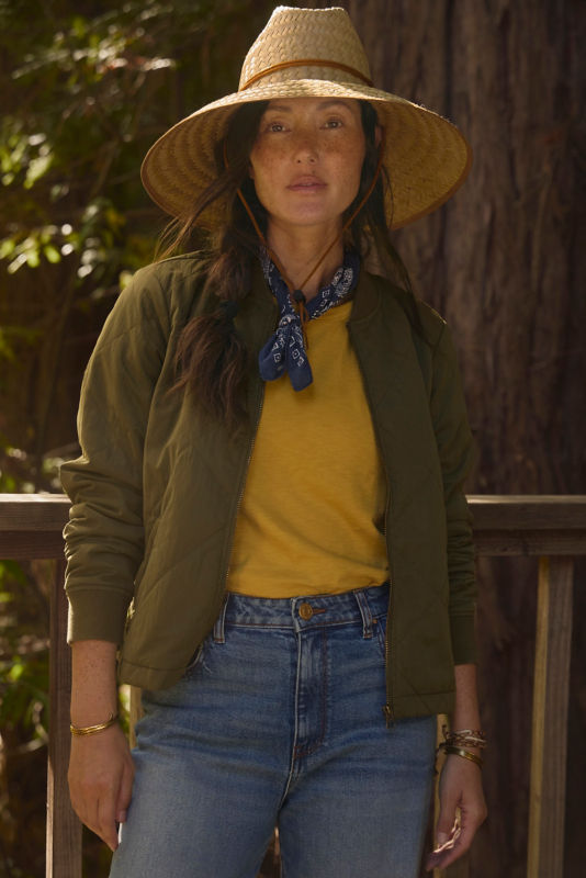 Woman standing in front of a tree wearing green jacket, yellow t-shirt, and jeans.