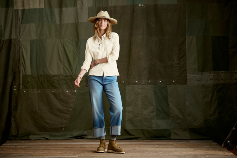 Woman wearing white button down, straw hat, and jeans in a studio.