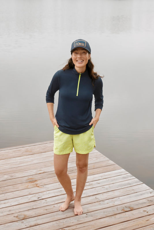 Woman in navy quarter-zip top, yellow shorts, and ballcap standing on a dock.