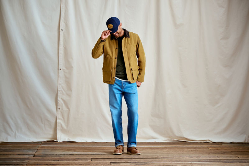 A man wearing a tan jacket with blue jeans and a navy ball cap.