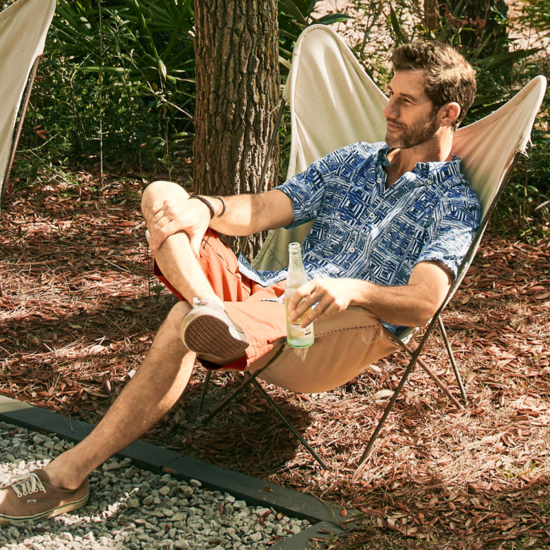 A man sits in a camp chair in the woods near a river holding a beverage.