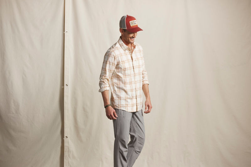 A model wearing a light plaid button-up shirt over gray pants with a red and a gray Orvis baseball cap.