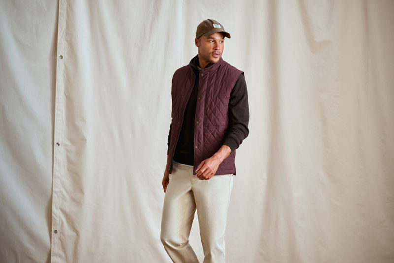 A man stands in front of a canvas wearing a maroon quilted vest over a brown sweater.