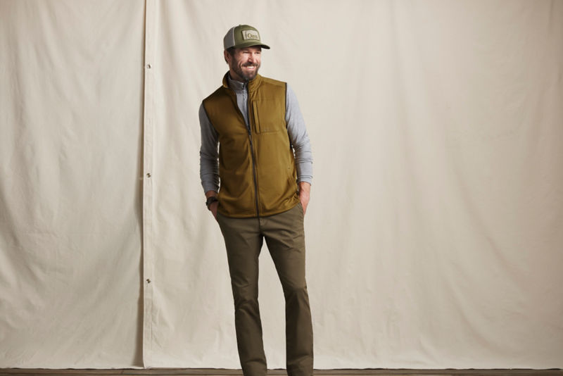 A model wearing an Orvis baseball hat and a gold vest over a grey shirt.