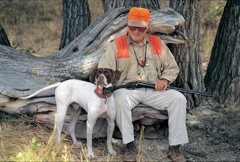 Leigh H Perkins in hunting gear, sitting on a tree stump next to a hunting dog.