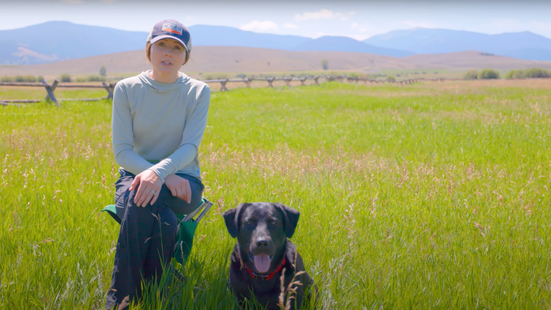 A woman and her panting dog sitting in the tall grass in an open field