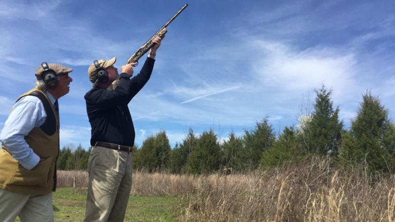 An instructor guiding another person shooting a shotgun into the air.