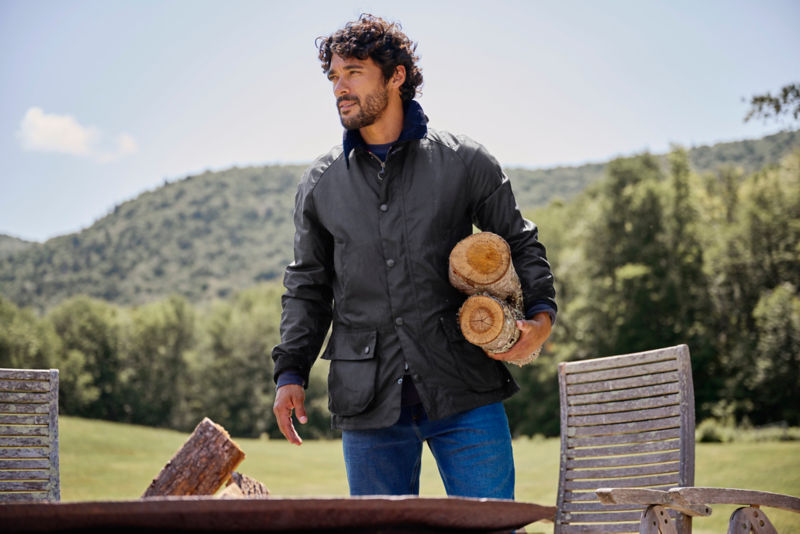 A man wearing a waxed Barbour jacket carries two logs under his arm.