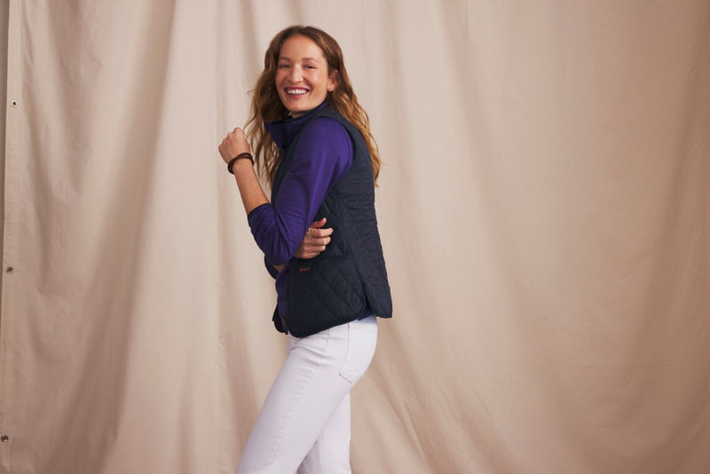 A smiling woman wears a black puffy vest over a purple fleece and white jeans.