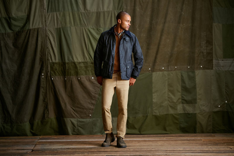 A model wearing a black jacket and khaki pants stands in front of a green canvas backdrop.