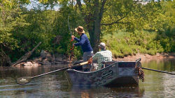 Two anglers in a boat; on casts into the river while the other rows.
