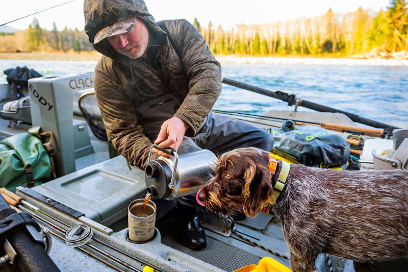A man pouring coffee into a mug while in a boat with his dog.