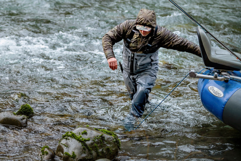 An angler wearing zip-front waders steadies themself against their raft as they wade in turbulent water