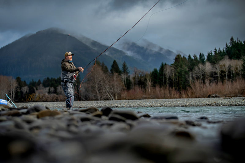 An angler dressed in full gear stands in a river while casting.