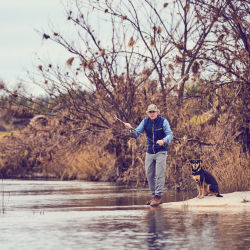A man fishes from the riverside with his watching dog