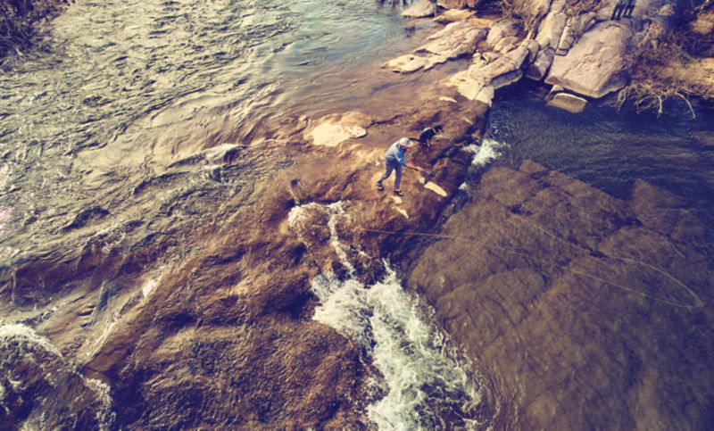 An angler casts from a dry bit of rock in a wide, shallow river.