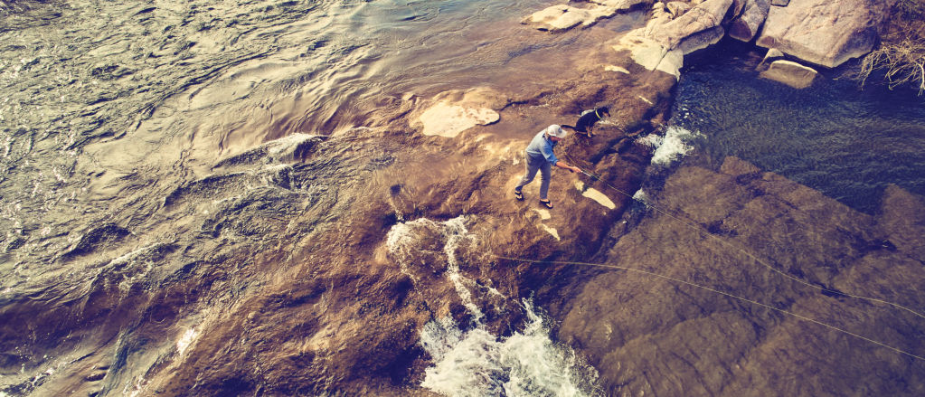 An angler casts from a dry bit of rock in a wide, shallow river.