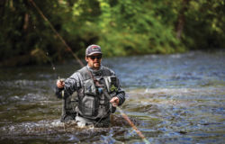 Jesse standing waist-deep in the river, wearing waders and casting a line.