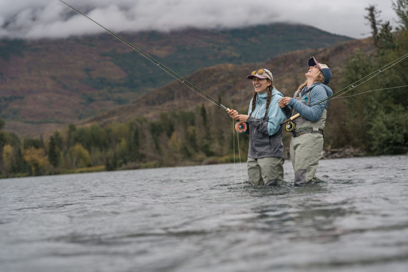 Two anglers in waders smiling and laughing in the river as they're casting together.
