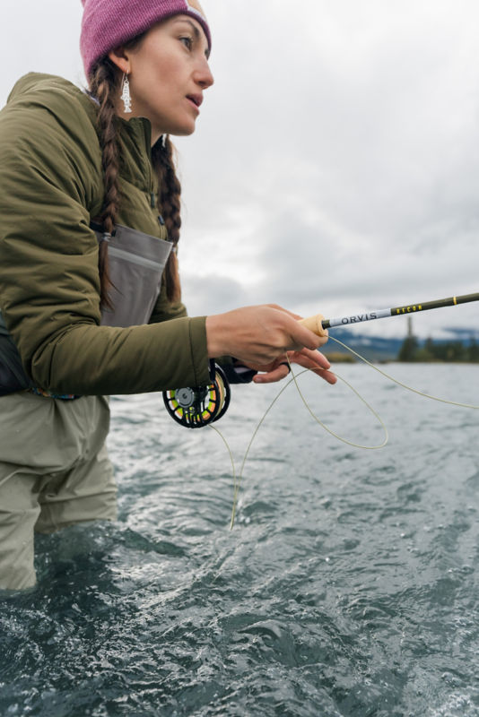 An angler leans into the cast from thigh-deep in fast-moving water.
