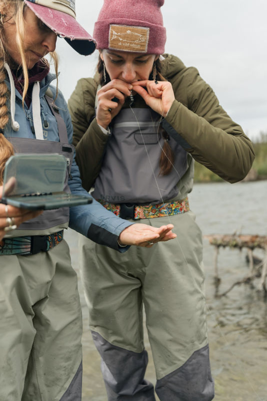 An angler tying a fly with her mouth while another angler watches. 
