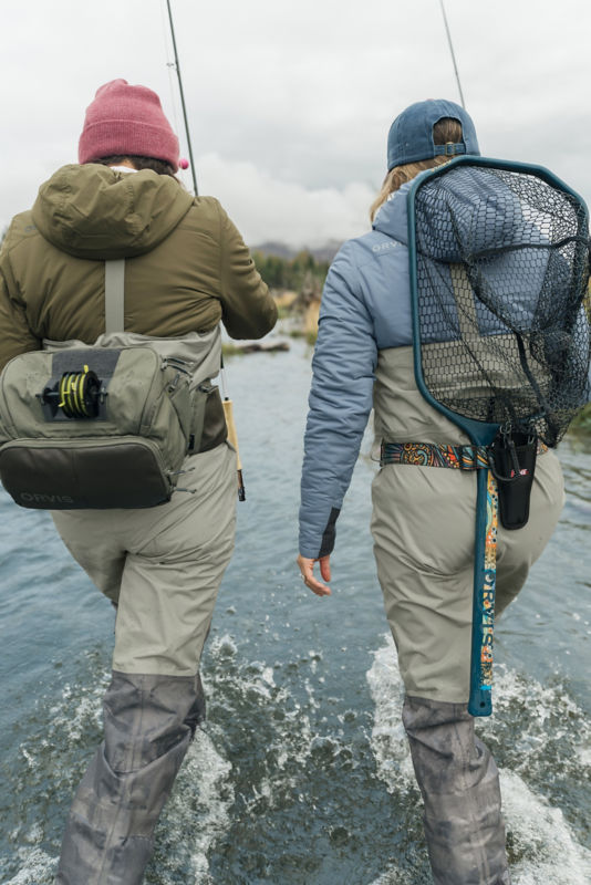 Two anglers from the back as they splash downstream with fishing packs and nets.