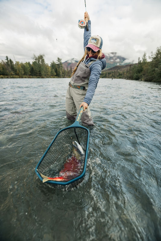 An angler grins as she lifts her fly rod high with one hand while she nets her fish with the other.
