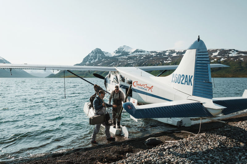 A small group of people unload fishing equipment from a small plane at the shore in Alaska.