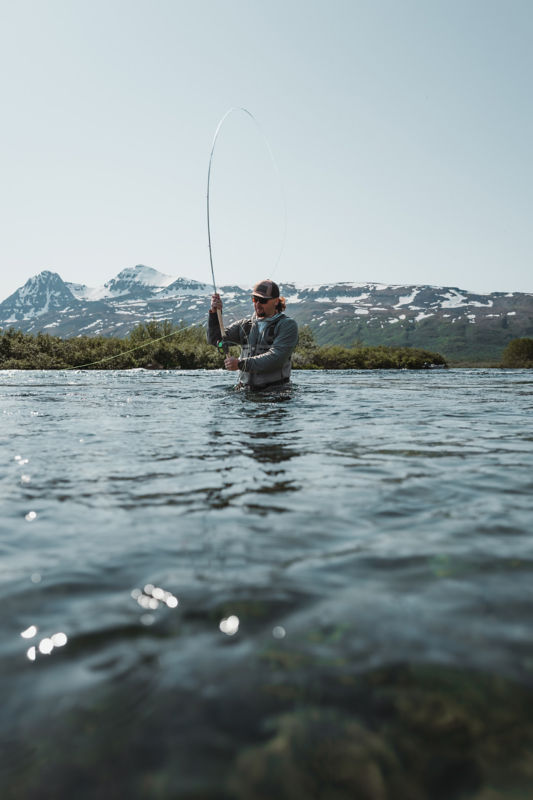 An angler pull his rod back for the cast from waist-deep in Alaskan waters with mountains in the background.