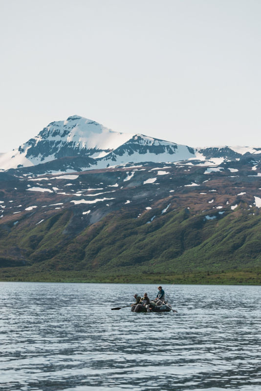A long-distance view of several people fishing from a raft under a snow-topped mountain.