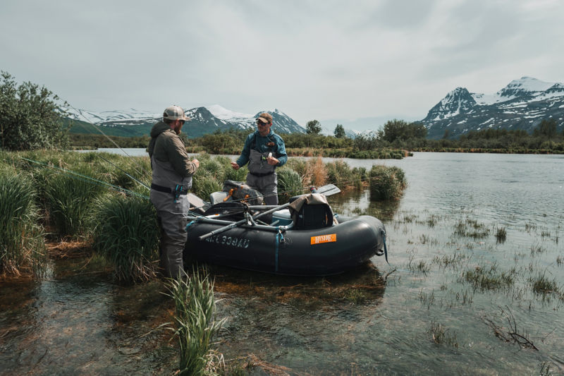 Two anglers gear up next to a raft with snowy mountains in the background.