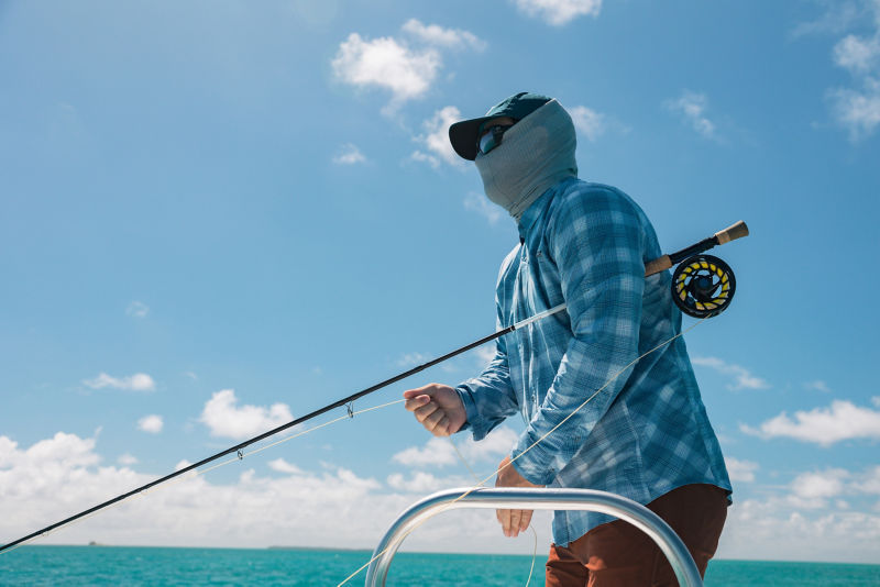 An angler wrapped in a buff and hat holds his fly rod under his arm.