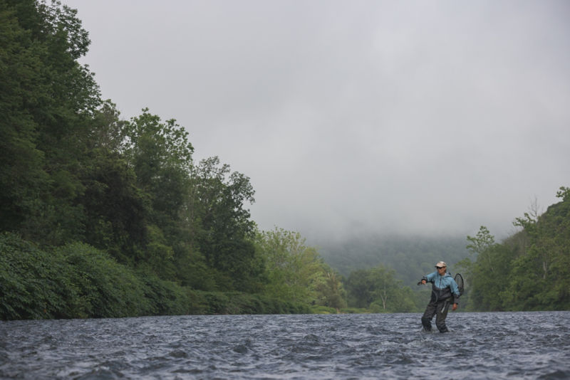 An angler pulls back her fly rod from shin-deep in a choppy river.