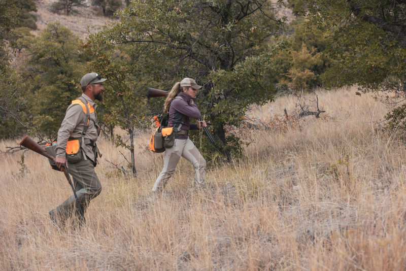 Two hunters wear blaze orange as they navigate through a fields of tall golden grass.