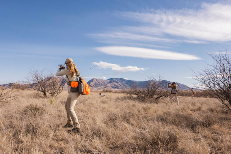 A hunter stands and raises her shotgun toward a bird in the distance.