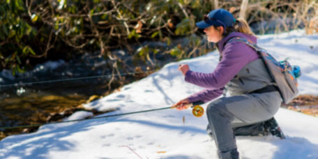An angler casts on one knee, riverside in the snow.