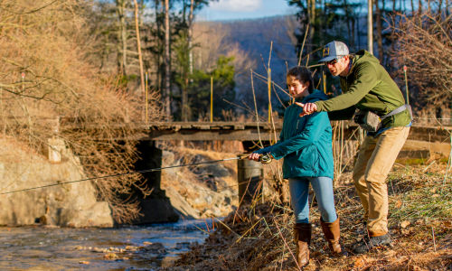 An angler and his daughter fish alongside a shallow river.
