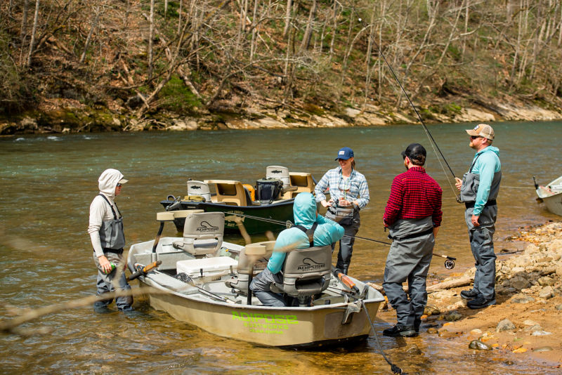 A group of anglers stand next to a boat, riverside.